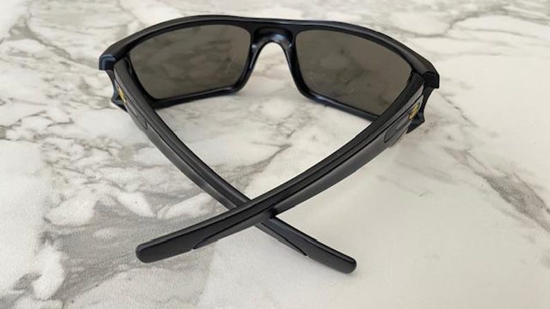 May include: Black sunglasses with dark lenses. The sunglasses have a rectangular frame and are positioned with the arms crossed. The lenses are a dark grey colour, and the frame is a matte black. The sunglasses are on a white marble surface.