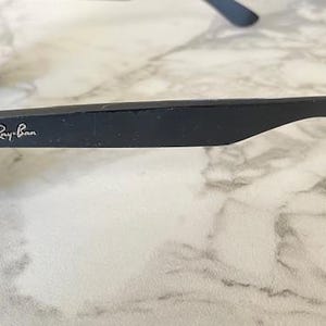 May include: Black sunglasses with the Ray-Ban logo on the temple. The sunglasses are set against a marble background. The frame is a matte black color.