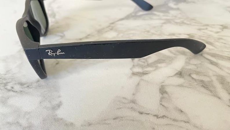 May include: Black sunglasses with the Ray-Ban logo on the temple. The sunglasses are set against a marble background. The frame is a matte black color.