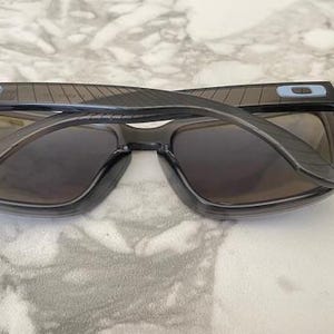 May include: Gray sunglasses with dark lenses, viewed from above. The frames are a translucent gray with a rectangular shape. Small blue accents are visible on the temples. The sunglasses are resting on a marble surface.