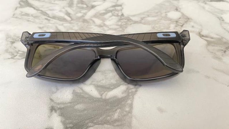 May include: Gray sunglasses with dark lenses, viewed from above. The frames are a translucent gray with a rectangular shape. Small blue accents are visible on the temples. The sunglasses are resting on a marble surface.