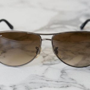 May include: Aviator-style sunglasses with a brown gradient lens and a dark metal frame. The Ray-Ban logo is visible on the upper left lens. The sunglasses have a classic teardrop shape.