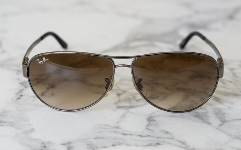 May include: Aviator-style sunglasses with a brown gradient lens and a dark metal frame. The Ray-Ban logo is visible on the upper left lens. The sunglasses have a classic teardrop shape.