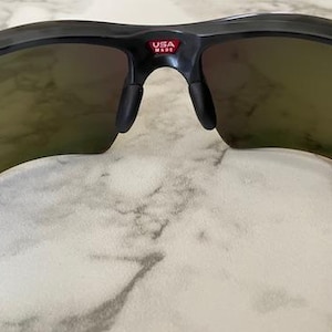 Puede incluir: Gafas de sol negras con lentes tintadas. Las gafas tienen un dise&ntilde;o curvo y un peque&ntilde;o logotipo "USA MADE" rojo y blanco en el puente. Las lentes tienen una apariencia reflectante y espejada.