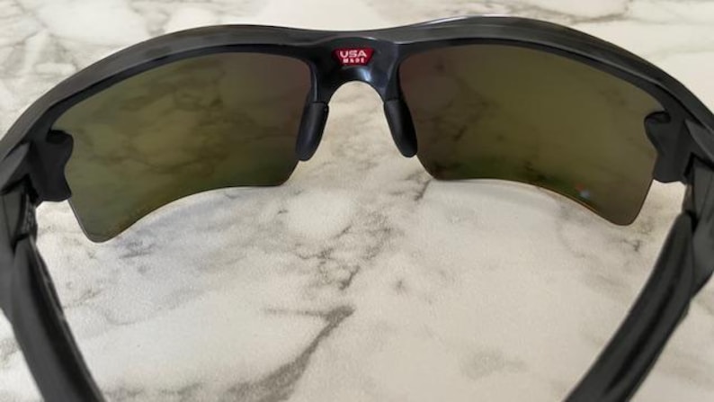 Puede incluir: Gafas de sol negras con lentes tintadas. Las gafas tienen un dise&ntilde;o curvo y un peque&ntilde;o logotipo "USA MADE" rojo y blanco en el puente. Las lentes tienen una apariencia reflectante y espejada.