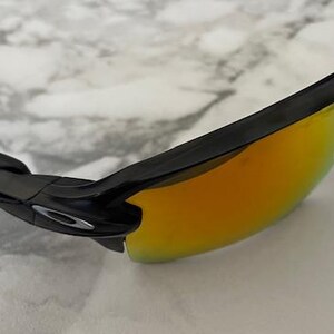 Puede incluir: Gafas de sol negras con lentes espejadas de color naranja-amarillo. Las gafas de sol tienen un dise&ntilde;o elegante y moderno con montura y patillas negras. Las lentes reflejan un color brillante y c&aacute;lido, y el logotipo de Oakley es visible.