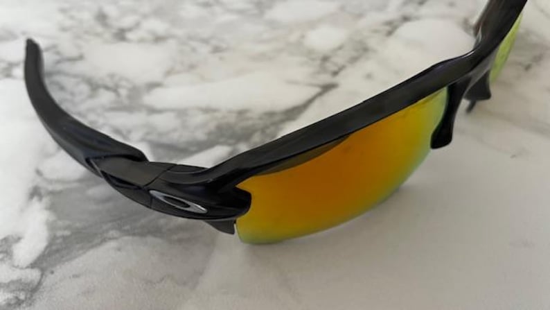 Puede incluir: Gafas de sol negras con lentes espejadas de color naranja-amarillo. Las gafas de sol tienen un dise&ntilde;o elegante y moderno con montura y patillas negras. Las lentes reflejan un color brillante y c&aacute;lido, y el logotipo de Oakley es visible.