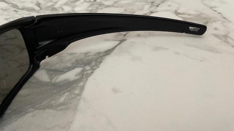 May include: Close-up of black sunglasses with a sleek, modern design. The sunglasses have a thick frame and arms, with a small rectangular detail at the temple. The lenses are dark, and the overall aesthetic is minimalist.