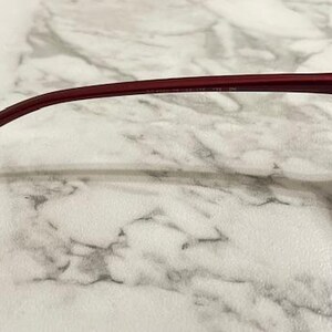 May include: Close-up of a pair of burgundy-colored sunglasses. The frame is a deep red hue, with a curved temple arm. The brand name "Hall & Blue" is printed on the temple. The lenses are a gradient color.
