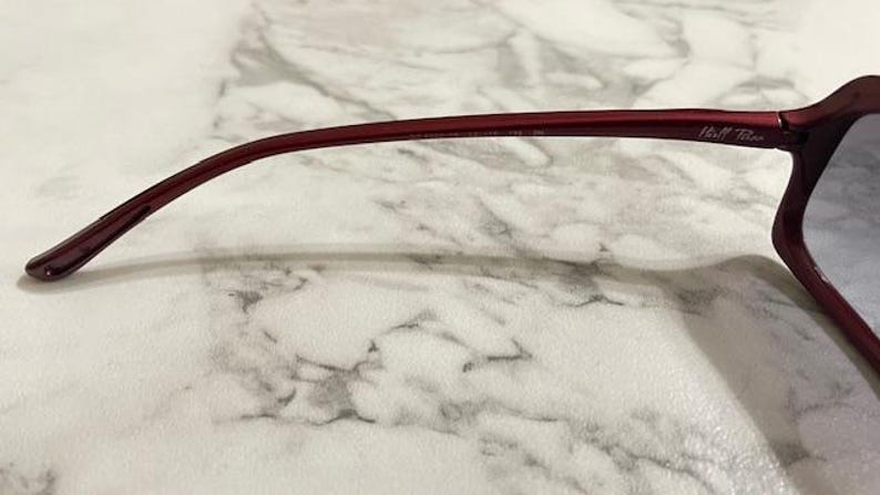 May include: Close-up of a pair of burgundy-colored sunglasses. The frame is a deep red hue, with a curved temple arm. The brand name "Hall & Blue" is printed on the temple. The lenses are a gradient color.