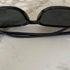 Puede incluir: Gafas de sol negras con lentes oscuras. Las monturas son de color negro brillante y las patillas son finas y curvadas. Las gafas de sol est&aacute;n sobre una superficie clara. Las lentes tienen forma ovalada.