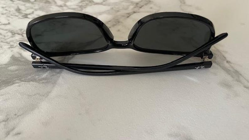 Puede incluir: Gafas de sol negras con lentes oscuras. Las monturas son de color negro brillante y las patillas son finas y curvadas. Las gafas de sol est&aacute;n sobre una superficie clara. Las lentes tienen forma ovalada.