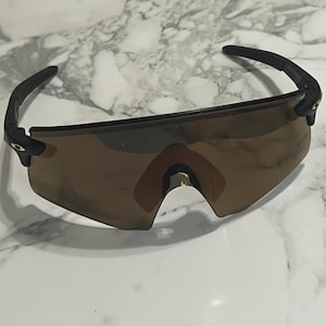 Puede incluir: Gafas de sol deportivas con montura negra y lentes marrones. Las gafas tienen un diseño elegante y moderno, con una barra de cejas y patillas curvadas. Las lentes son de color marrón degradado.