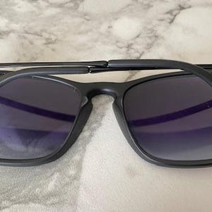 May include: Black sunglasses with dark lenses. The sunglasses have a square frame and a matte finish. The arms are folded, revealing the inside of the lenses. The Ray-Ban logo is visible on the temples.