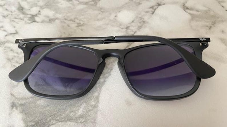 May include: Black sunglasses with dark lenses. The sunglasses have a square frame and a matte finish. The arms are folded, revealing the inside of the lenses. The Ray-Ban logo is visible on the temples.