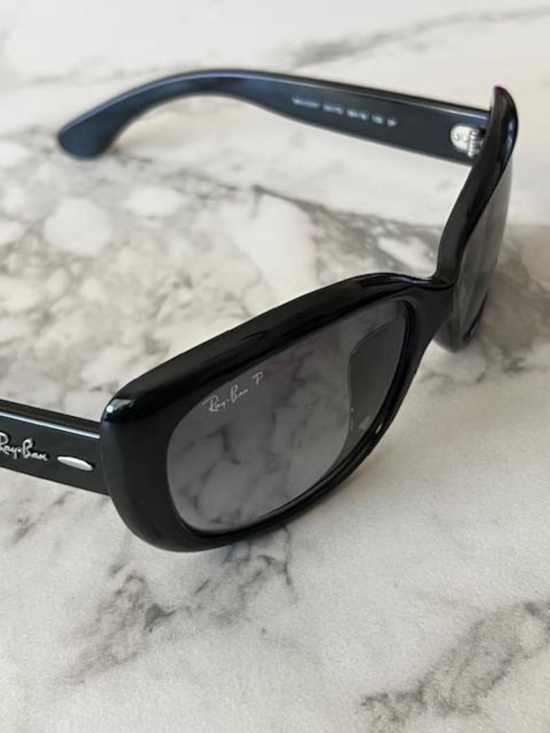 K&ouml;nnte beinhalten: Schwarze Ray-Ban Sonnenbrille mit rechteckigem Rahmen und Farbverlaufsgl&auml;sern. Das Ray-Ban-Logo ist auf dem B&uuml;gel und der Linse zu sehen. Die Sonnenbrille liegt auf einem Marmorhintergrund.