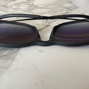 May include: Black sunglasses with a unique, angular frame design. The lenses are a dark, gradient color, and the arms are thin and sleek. The sunglasses are set against a light, marble-like background.