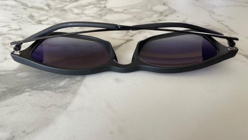 May include: Black sunglasses with a unique, angular frame design. The lenses are a dark, gradient color, and the arms are thin and sleek. The sunglasses are set against a light, marble-like background.