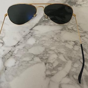 May include: Aviator-style sunglasses with a gold metal frame and dark lenses. The sunglasses have a double bridge and black earpieces. The lenses are a dark color, and the frame is a shiny gold color.