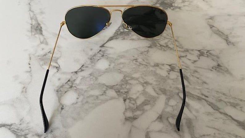 May include: Aviator-style sunglasses with a gold metal frame and dark lenses. The sunglasses have a double bridge and black earpieces. The lenses are a dark color, and the frame is a shiny gold color.