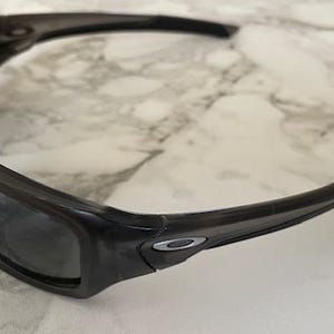 May include: Dark gray sunglasses with a wraparound design. The sunglasses have a thick, rectangular frame and dark lenses. The Oakley logo is visible on the temple. The sunglasses are designed for sports and outdoor activities.