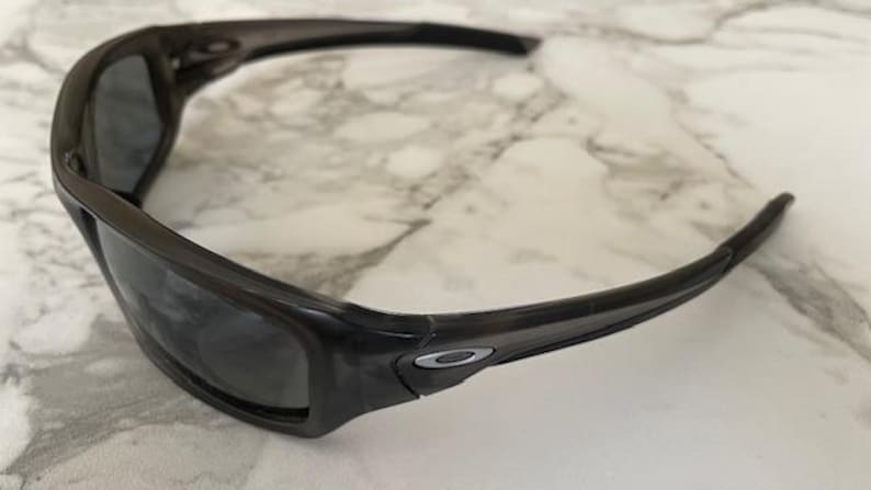 May include: Dark gray sunglasses with a wraparound design. The sunglasses have a thick, rectangular frame and dark lenses. The Oakley logo is visible on the temple. The sunglasses are designed for sports and outdoor activities.