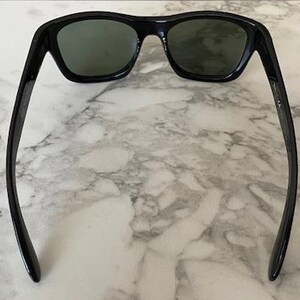 Puede incluir: Gafas de sol negras con lentes verde oscuro. Las gafas tienen una montura rectangular y patillas curvas. La imagen est&aacute; tomada desde arriba, mostrando las gafas sobre un fondo de m&aacute;rmol.