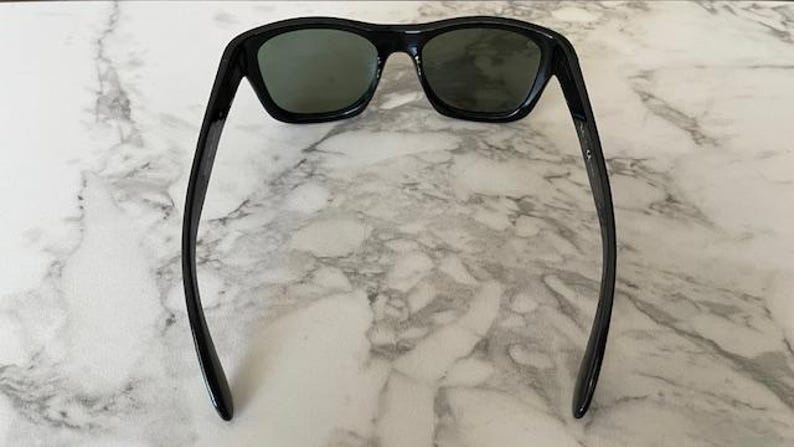 Puede incluir: Gafas de sol negras con lentes verde oscuro. Las gafas tienen una montura rectangular y patillas curvas. La imagen est&aacute; tomada desde arriba, mostrando las gafas sobre un fondo de m&aacute;rmol.