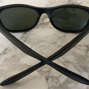 May include: Black sunglasses with dark green lenses. The arms of the sunglasses are crossed, showing the back of the frame. The sunglasses are on a white and gray marble surface.
