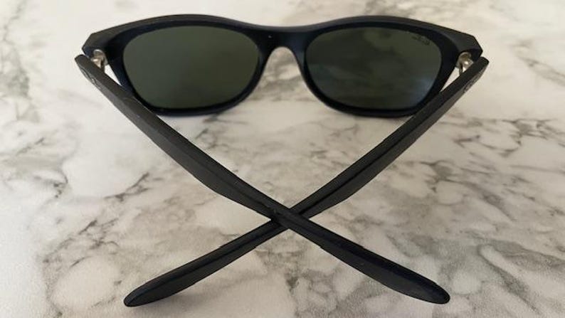 May include: Black sunglasses with dark green lenses. The arms of the sunglasses are crossed, showing the back of the frame. The sunglasses are on a white and gray marble surface.