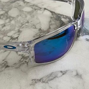 May include: Clear-framed sunglasses with blue mirrored lenses. The sunglasses have a sleek, modern design with a subtle blue logo on the temple. The lenses reflect a vibrant blue hue, offering a stylish and functional accessory.