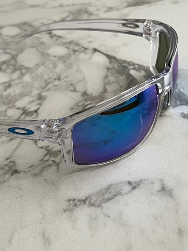 May include: Clear-framed sunglasses with blue mirrored lenses. The sunglasses have a sleek, modern design with a subtle blue logo on the temple. The lenses reflect a vibrant blue hue, offering a stylish and functional accessory.