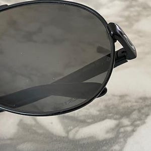 May include: Black aviator-style sunglasses with dark tinted lenses. The sunglasses have a sleek, modern design with a thin black frame and a double bridge. The lenses are oval-shaped and provide full coverage.