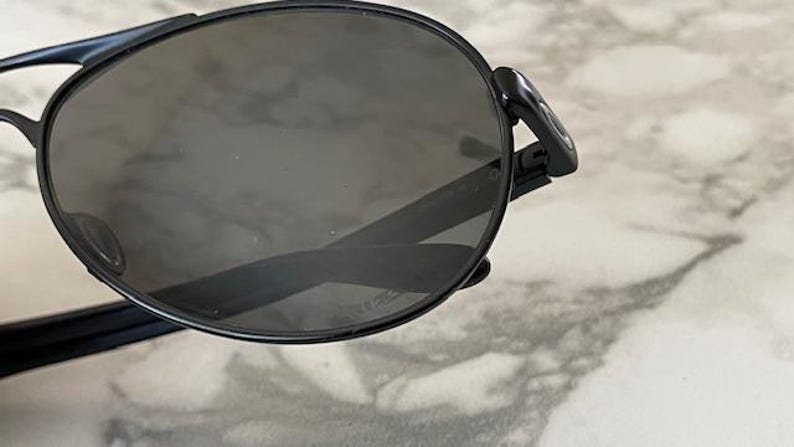 May include: Black aviator-style sunglasses with dark tinted lenses. The sunglasses have a sleek, modern design with a thin black frame and a double bridge. The lenses are oval-shaped and provide full coverage.