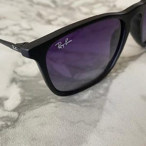 May include: Black sunglasses with purple gradient lenses. The Ray-Ban logo is visible on the upper left lens. The sunglasses are set against a white and gray marble background.