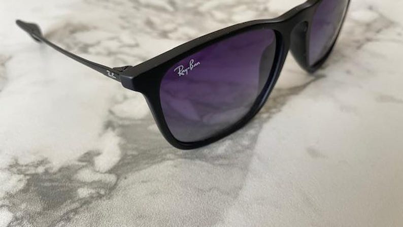 May include: Black sunglasses with purple gradient lenses. The Ray-Ban logo is visible on the upper left lens. The sunglasses are set against a white and gray marble background.