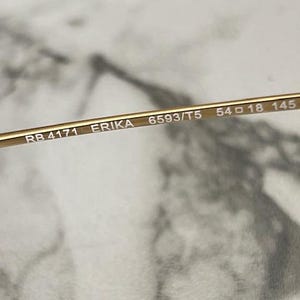 May include: Close-up of a gold-colored eyeglass frame. The text "RB 4171 ERIKA 6593/T5 54 18 145 3P" is printed on the frame. The frame has a sleek, modern design. The lens width is 54 mm.