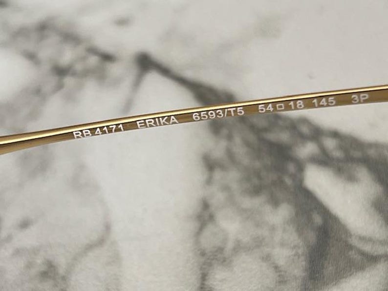 May include: Close-up of a gold-colored eyeglass frame. The text "RB 4171 ERIKA 6593/T5 54 18 145 3P" is printed on the frame. The frame has a sleek, modern design. The lens width is 54 mm.