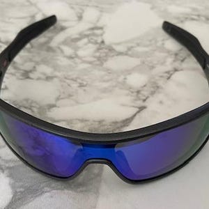 Może przedstawiać: Czarne okulary przeciwsłoneczne z niebieskimi lustrzanymi soczewkami. Okulary mają elegancki, sportowy design z zakrzywioną oprawką. Soczewki odbijają żywy niebieski kolor, a oprawka jest matowa czarna.