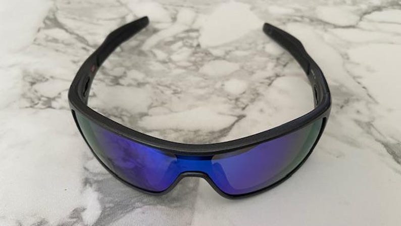 Może przedstawiać: Czarne okulary przeciwsłoneczne z niebieskimi lustrzanymi soczewkami. Okulary mają elegancki, sportowy design z zakrzywioną oprawką. Soczewki odbijają żywy niebieski kolor, a oprawka jest matowa czarna.