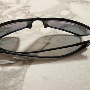 May include: Black sunglasses with gray lenses are folded on a marble surface. The sunglasses have a sleek, modern design with a matte finish. The temples are thin and connect to the frame with silver accents. The lenses are tinted.