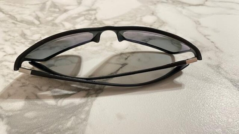May include: Black sunglasses with gray lenses are folded on a marble surface. The sunglasses have a sleek, modern design with a matte finish. The temples are thin and connect to the frame with silver accents. The lenses are tinted.