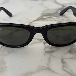 K&ouml;nnte beinhalten: Schwarze Sonnenbrille mit klassischem, eckigem Rahmen. Die Gl&auml;ser sind dunkel, und das Ray-Ban-Logo ist auf dem rechten Glas zu sehen. Die B&uuml;gel sind dick und biegen sich nach unten, um &uuml;ber die Ohren zu passen.