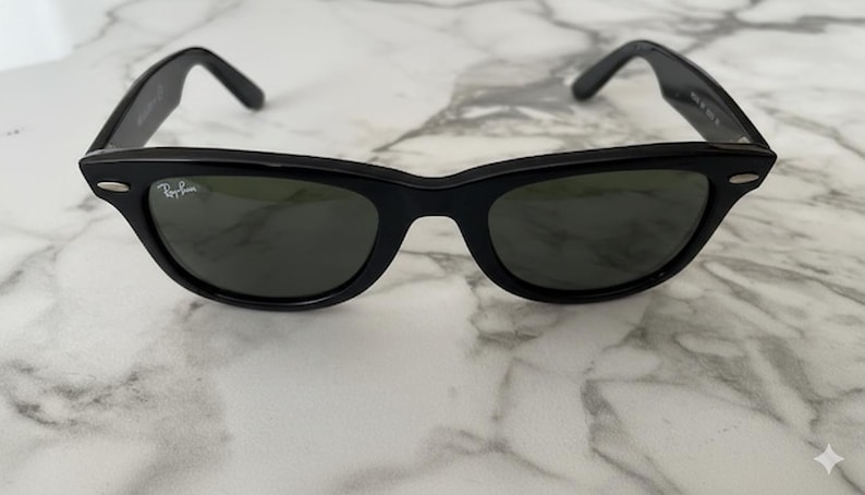 K&ouml;nnte beinhalten: Schwarze Sonnenbrille mit klassischem, eckigem Rahmen. Die Gl&auml;ser sind dunkel, und das Ray-Ban-Logo ist auf dem rechten Glas zu sehen. Die B&uuml;gel sind dick und biegen sich nach unten, um &uuml;ber die Ohren zu passen.
