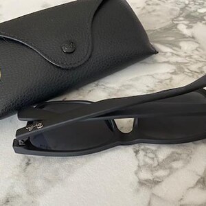 Pu&ograve; includere: Occhiali da sole neri e custodia nera abbinata. Gli occhiali sono piegati e hanno un design elegante. La custodia &egrave; realizzata in materiale nero strutturato e presenta un logo dorato. Su una superficie di marmo.
