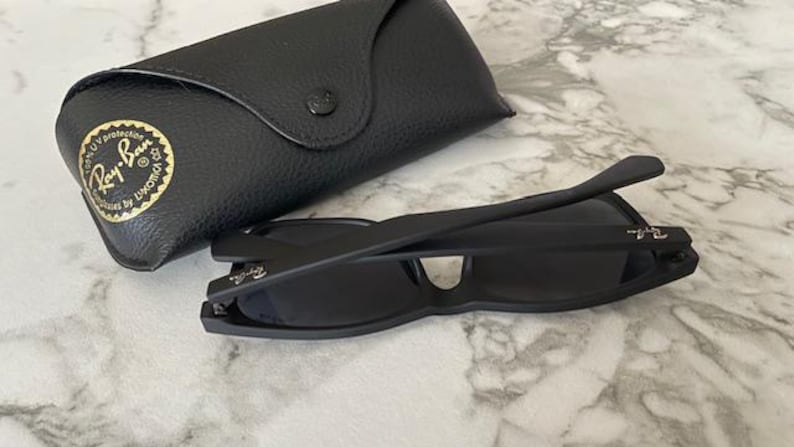 Pu&ograve; includere: Occhiali da sole neri e custodia nera abbinata. Gli occhiali sono piegati e hanno un design elegante. La custodia &egrave; realizzata in materiale nero strutturato e presenta un logo dorato. Su una superficie di marmo.