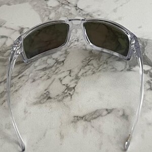 May include: Clear-framed sunglasses with dark lenses. The sunglasses have a rectangular shape and a transparent frame. The lenses appear to be a dark shade, providing protection from the sun. The arms of the sunglasses are also clear.