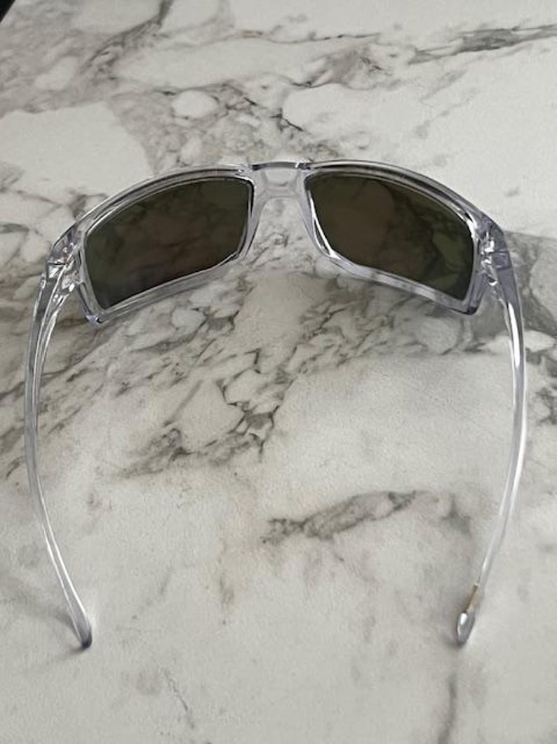 May include: Clear-framed sunglasses with dark lenses. The sunglasses have a rectangular shape and a transparent frame. The lenses appear to be a dark shade, providing protection from the sun. The arms of the sunglasses are also clear.