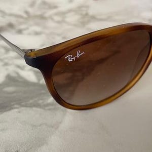 May include: Brown sunglasses with a tortoiseshell frame and gradient lenses. The Ray-Ban logo is visible on the lens. The sunglasses have a classic, rounded shape and metal arms.