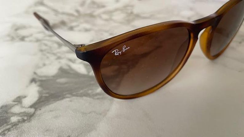 May include: Brown sunglasses with a tortoiseshell frame and gradient lenses. The Ray-Ban logo is visible on the lens. The sunglasses have a classic, rounded shape and metal arms.